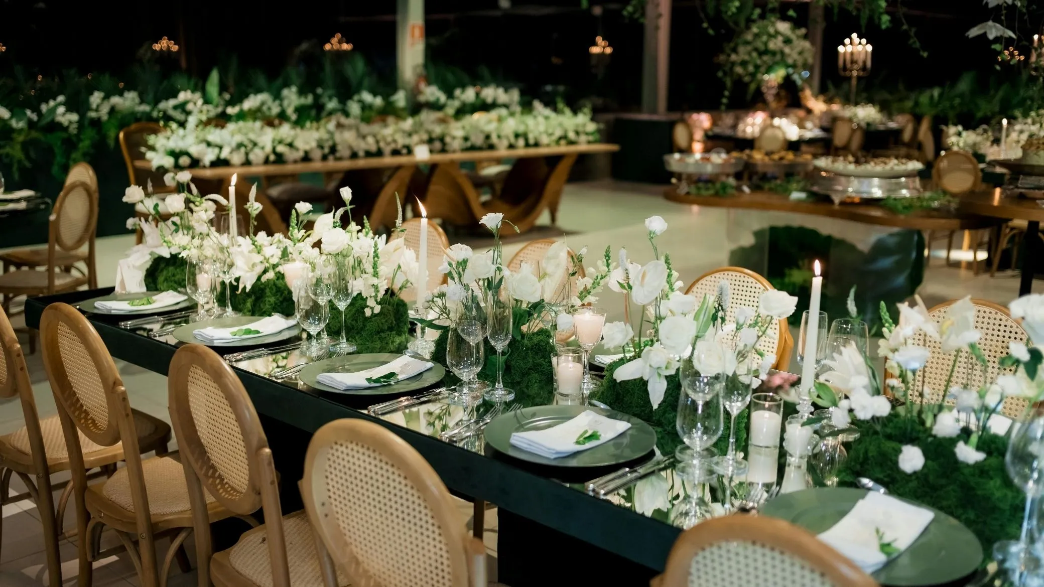 Longue table de réception sombre dressée de fleurs blanches, de mousse verte et de bougies, entourée de chaises cannées, dans une salle éclairée le soir