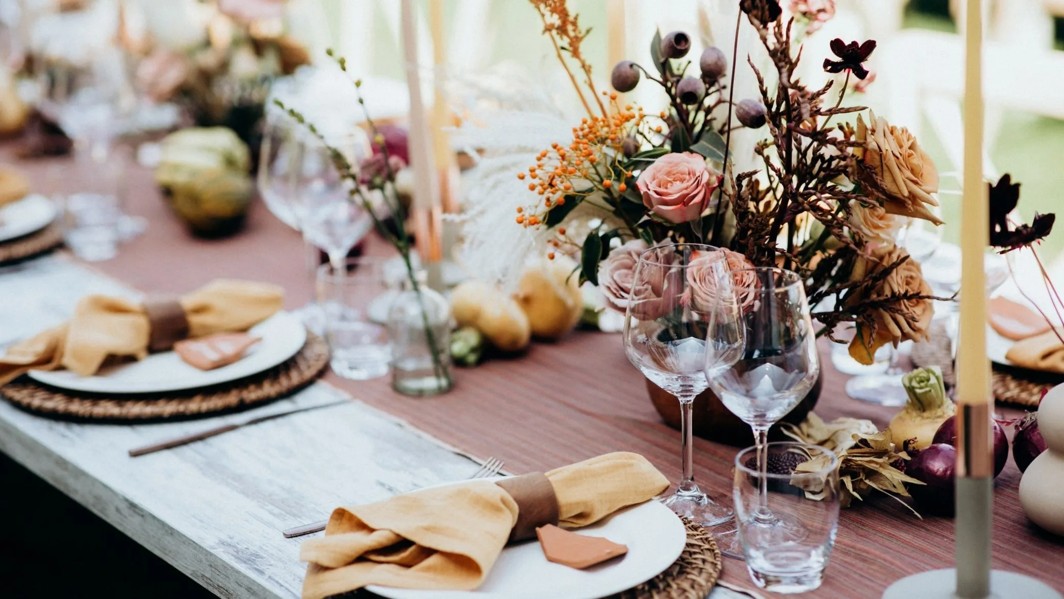 Table de mariage en bois clair avec un chemin de table rose, des serviettes ocre nouées d'un anneau de cuir, de petits marque-places en terre cuite et un bouquet de roses et de baies